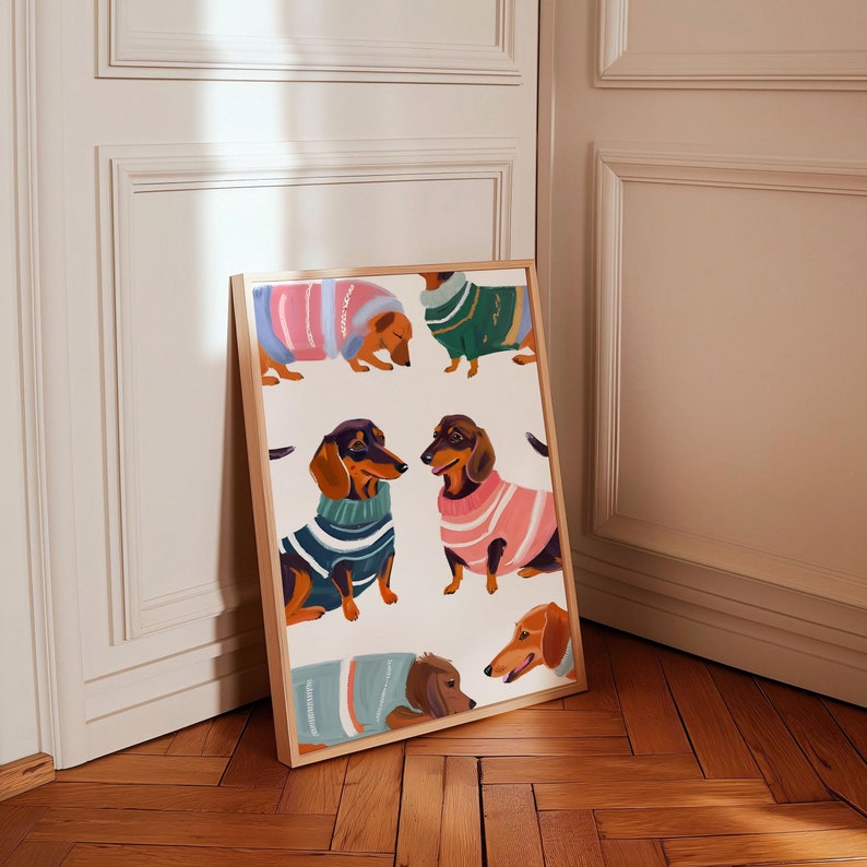 May include: A framed print featuring a whimsical illustration of five dachshund dogs wearing colorful sweaters. The dogs are depicted in various poses, with some looking directly at the viewer and others looking away. The background is a light cream color.