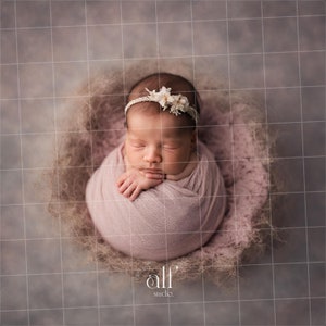 May include: A newborn baby girl is sleeping peacefully in a pink swaddle, wearing a white flower headband. The baby is lying in a nest-like basket with a light brown background.