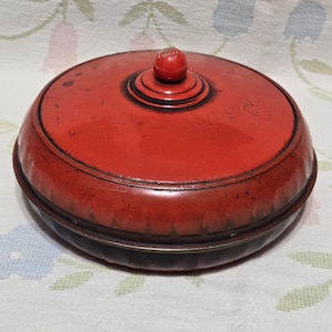 May include: A round, red lidded container with a smooth, glossy finish and a small, centered knob handle on top. The lid features concentric circular ridges around the knob, and the base has a slightly darker, muted red tone with subtle surface wear.