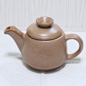 May include: A small, speckled brown ceramic teapot with a matching lid and a rounded handle. The teapot has a spout for pouring and a small knob on the lid. The teapot is designed for brewing tea.