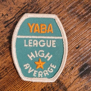 May include: A teal blue embroidered patch with white stitching. The patch has the text "YABA LEAGUE HIGH AVERAGE" in white with a yellow star in the center.