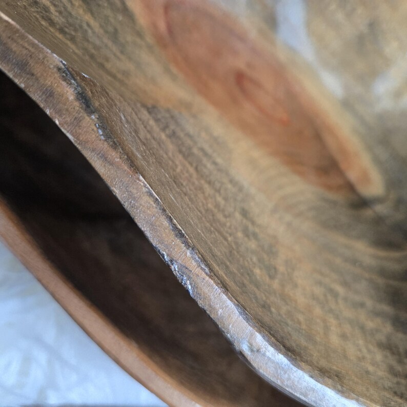 May include: Close-up of the edge of a large wooden bowl. The bowl is made of light brown wood with darker brown streaks. The edge of the bowl is slightly chipped.