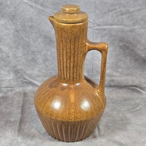 May include: A brown ceramic pitcher with a lid. The pitcher has a rounded base and a tall, ribbed body. It features a handle and a spout for pouring. The lid is round and fits snugly on top. The pitcher has a speckled, textured surface.