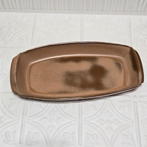 May include: A rectangular, brown ceramic serving platter with rounded corners. The platter has a speckled texture and a slightly raised rim. The background is a white, patterned surface.