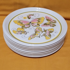 Pu&ograve; includere: Un set di dieci piatti da cena in ceramica bianca con un design a farfalla. Le farfalle sono gialle, rosa e verdi con contorni neri. I piatti hanno un bordo verde e giallo.