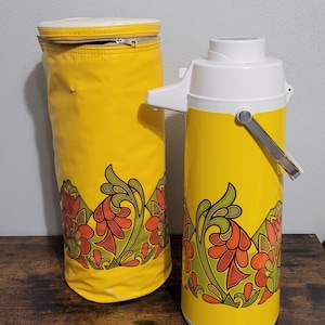 May include: A yellow and white vintage thermal carafe with a floral design and a matching yellow carrying case. The carafe has a white lid and a silver handle.