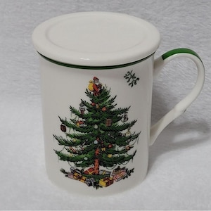 Op de afbeelding: Een witte keramische mok met een groene handgreep en rand, met een kerstboomontwerp met de kerstman erop. De mok heeft een bijpassende deksel. Cadeaus staan aan de voet van de boom.