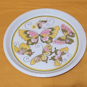 Pu&ograve; includere: Un vassoio da portata in ceramica bianca con un design decorativo di farfalle rosa, gialle e verdi. Le farfalle hanno motivi intricati e sono disposte in un motivo circolare sul vassoio.