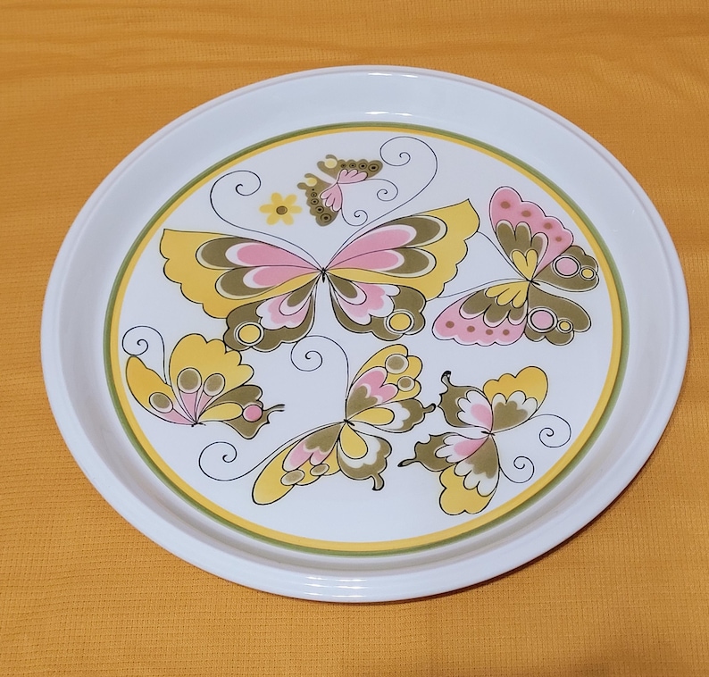 Pu&ograve; includere: Un vassoio da portata in ceramica bianca con un design decorativo di farfalle rosa, gialle e verdi. Le farfalle hanno motivi intricati e sono disposte in un motivo circolare sul vassoio.