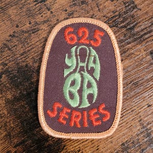 May include: A brown fabric patch with orange stitching. The patch has the text "625 YA BA SERIES" in green and orange embroidery.