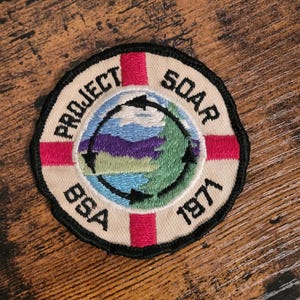 Può includere: Una toppa rotonda bianca con una croce rossa e il testo "PROJECT SOAR BSA 1971". Il centro della toppa presenta un paesaggio ricamato colorato con un cielo blu, alberi verdi, montagne viola e un lago blu.