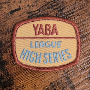 Pode incluir: Um remendo bordado amarelo com uma borda marrom. O remendo diz "YABA LEAGUE HIGH SERIES" em fio azul.