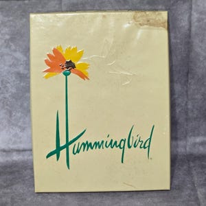 Puede incluir: Una caja crema con un diseño floral y una silueta de colibrí. La palabra "Hummingbird" está escrita en verde con letra cursiva. La flor tiene pétalos naranjas y amarillos.