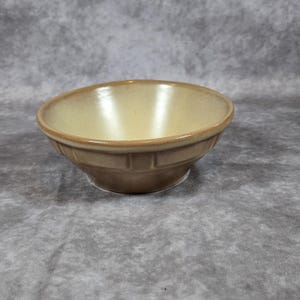 Puede incluir: Un cuenco de cerámica marrón con interior amarillo claro. El cuenco tiene forma redondeada con una banda decorativa alrededor del centro. El fondo es gris moteado.