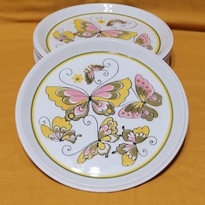 Pu&ograve; includere: Set di otto piatti da portata in ceramica bianca con un design a farfalla. Le farfalle sono gialle, rosa e verdi con contorni neri. I piatti hanno un bordo verde e giallo.