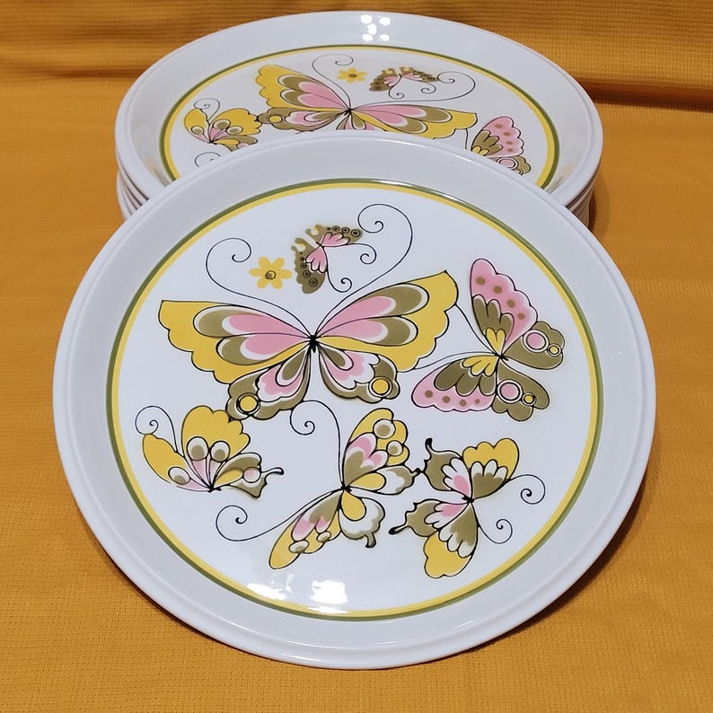 Pu&ograve; includere: Set di otto piatti da portata in ceramica bianca con un design a farfalla. Le farfalle sono gialle, rosa e verdi con contorni neri. I piatti hanno un bordo verde e giallo.