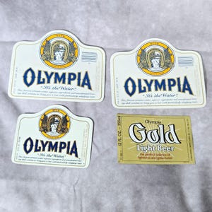 May include: Four Olympia beer labels. Three are white with blue text and a gold emblem. The text reads "Olympia" and "It's the Water." The fourth label is gold with black text that reads "Olympia Gold Light Beer."