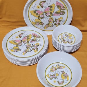 Pu&ograve; includere: Un set di stoviglie in ceramica bianca con un design a farfalla. Il set comprende un piatto da portata grande, piatti da insalata e ciotole. Le farfalle sono gialle, rosa e verdi.