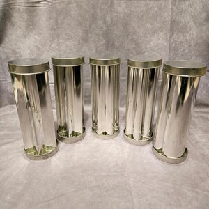 May include: Five shiny, cylindrical metal molds with fluted sides and flat lids. The molds are arranged in a row, each with a circular base. The metal has a reflective, silver appearance.