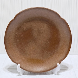 May include: A round, brown ceramic plate with a scalloped edge. The plate has a speckled texture and a matte finish. The plate is displayed on a white stand, showcasing its unique shape and colour.