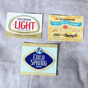 May include: Three vintage Cold Spring beer labels. One label reads "Cold Spring Light Strong Beer" with a red and gold design. Another label says "Cold Spring Export" with a blue and gold design. The third label says "Cold Spring Strong Beer".