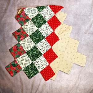 May include: A quilted Christmas tree-shaped table runner. The runner features a patchwork design with red, green, and white squares. The squares are patterned with floral and geometric designs. The backing is a light yellow fabric with small floral accents.