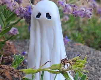 May include: A white ghost figurine with black eyes, standing in a garden setting with green plants and purple flowers.