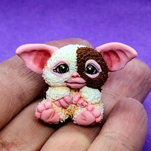 May include: A small, handmade figurine of a brown and white furry creature with pink ears and a pink nose. The creature has large, brown eyes and is sitting with its paws tucked under its body.