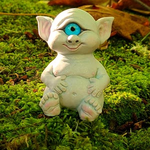 May include: A white, one-eyed, gnome-like figurine with a blue eye and a wide smile, sitting on a bed of green moss.