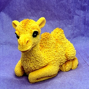 May include: A yellow camel figurine made of fondant. The camel is sitting on its haunches with its head turned to the side. The camel has a textured surface that looks like fur.