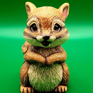 May include: A detailed figurine of a squirrel, featuring brown and cream fur, large brown eyes, and a friendly expression. The squirrel is posed with its paws together, set against a solid green background. This decorative item is likely a collectible or home decor piece.