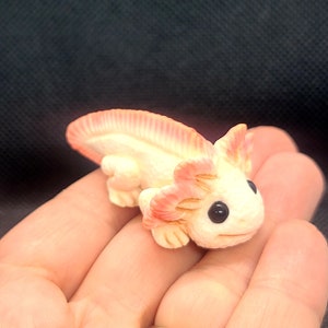 May include: A small, white axolotl figurine with pink gills and black eyes. The figurine is made of polymer clay and is about 3 inches long.