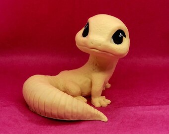 May include: A small, light beige ceramic gecko figurine with large black eyes. The gecko is sitting on its hind legs with its tail curled around its body.