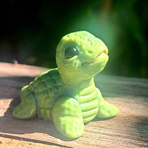 May include: A small green turtle figurine with a textured shell and large, black eyes. The turtle is sitting on a wooden surface.