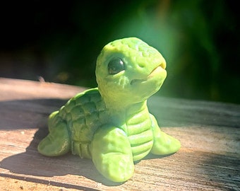 May include: A small green turtle figurine with a textured shell and large, black eyes. The turtle is sitting on a wooden surface.