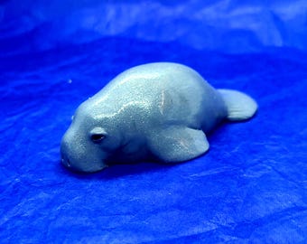May include: A light blue manatee figurine with a glittery finish. The figurine is lying on a blue background.