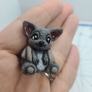 May include: A small gray and white bat figurine with big eyes and a cute expression. The bat is sitting with its wings folded and has a white belly.