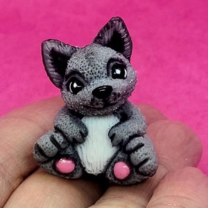 May include: A small gray and white ceramic figurine of a wolf pup sitting with its paws tucked in. The figurine has pink accents on its paws.