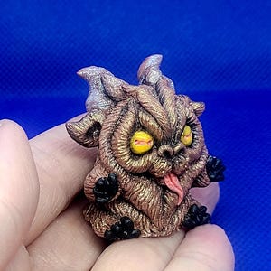 K&ouml;nnte beinhalten: Eine kleine, braune, pelzige Kreatur mit H&ouml;rnern, gelben Augen und einer herausgestreckten rosa Zunge. Die Kreatur ist aus Polymer Clay gefertigt und hat eine glitzernde Oberfl&auml;che.