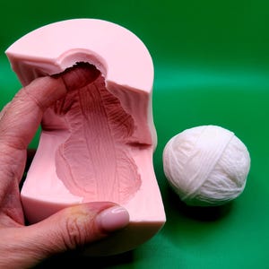 May include: A light pink silicone mold with a detailed interior design, held by a hand, next to a ball of white yarn. The mold has a curved top and a textured surface. The background is a solid green.