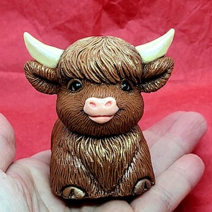 May include: A small, brown, fluffy Highland cow figurine with gold accents. The cow has a pink nose and is sitting on its hind legs.