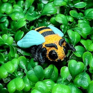 Op de afbeelding: Gedetailleerd bijenfiguur met geel-zwarte strepen, lichtblauwe vleugels en zwarte poten en antennes. De bij staat tegen een achtergrond van levendige groene bladeren, wat een naturalistische en kleurrijke scène creëert. Het figuur is een decoratief item.
