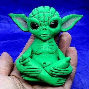 May include: A green ceramic figurine of a character from the Star Wars franchise, sitting in a meditative pose with hands clasped in front of them. The figurine has large eyes and a detailed face.