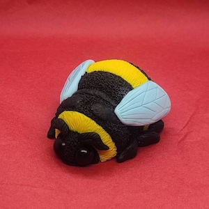 May include: A black and yellow bumble bee figurine with blue wings. The bee is lying on its side with its head facing the camera.