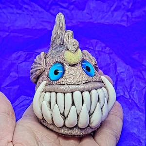 May include: A small ceramic figurine of a cartoon fish with a toothy grin. The fish is brown with blue eyes and a yellow fin. It has a row of white teeth and a textured surface.