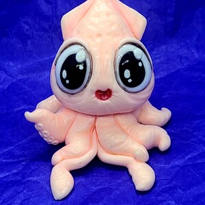 May include: A pink and white squid figurine with large, cartoon eyes. The squid has a small, red mouth and is sitting on a blue surface.