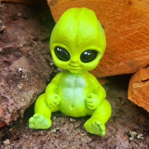 May include: A small green alien figurine with large eyes and a wide smile. The figurine is sitting on a piece of tree bark.