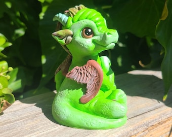 May include: A small green dragon figurine with brown wings, sitting with its wings folded. The dragon has a light green horn and large, black eyes.