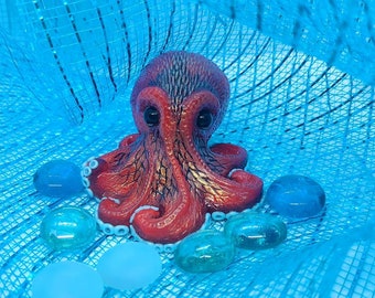 May include: A red and white octopus figurine with a textured surface, surrounded by blue glass marbles on a blue mesh background.
