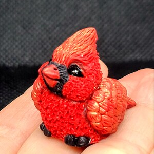 Puede incluir: Una pequeña figurita de cardenal rojo con ojos negros y pico negro. La figurita está hecha de un material texturizado que se asemeja a plumas.